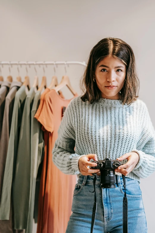 a woman holding a camera in front of a rack of clothes, by Nicolette Macnamara, wearing casual sweater, gemma chen, commercially ready, wearing a cute top