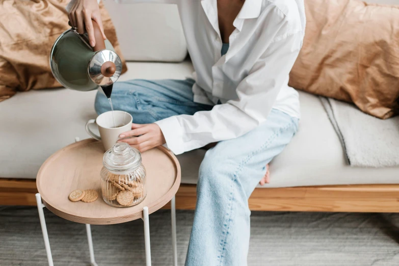 a woman sitting on a couch pouring a cup of coffee, by Emma Andijewska, trending on pexels, wearing a linen shirt, watering can, 2 - minute tabletop, snacks