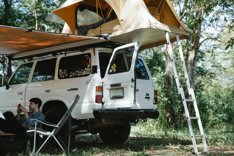 a man sitting under a tent on top of a white van, unsplash, ladders, jungle setting, brown, 1 6 x 1 6