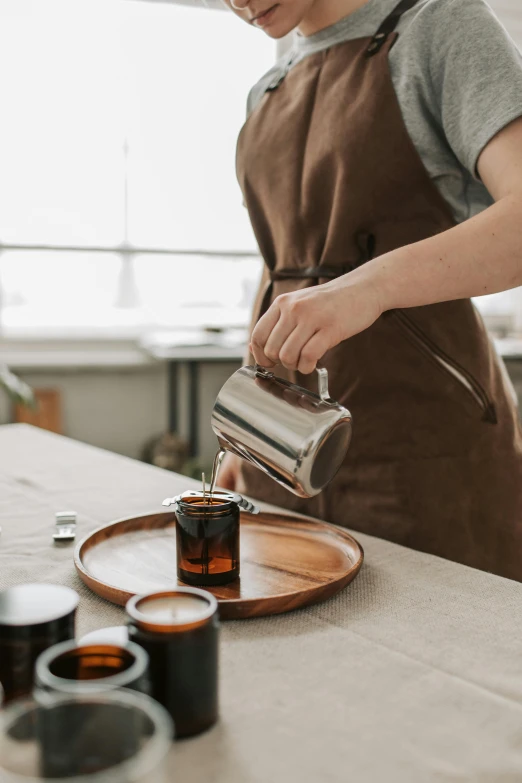 a woman in an apron pours a cup of coffee, by Frederik Vermehren, trending on unsplash, flasks, wax, on a table, manuka