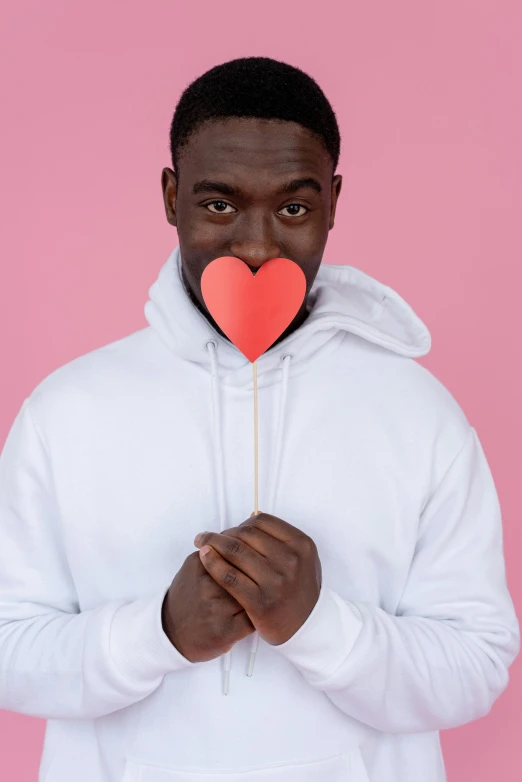 a man holding a paper heart in front of his face, an album cover, trending on pexels, romanticism, wearing a pastel pink hoodie, ( ( dark skin ) ), solid background, wearing white shirt