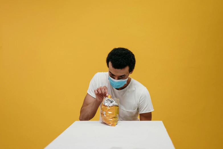 a man sitting at a table with a bag of doughnuts, by Adam Marczyński, pexels contest winner, yellow backdrop, head in a jar, medical mask, profile image