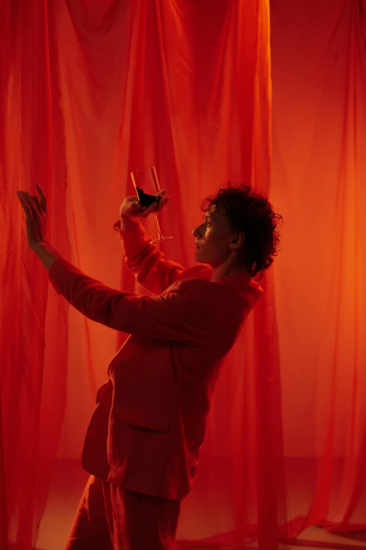 a man standing in front of a red curtain holding a wine glass, finn wolfhard, dancer, light red and orange mood, female magician