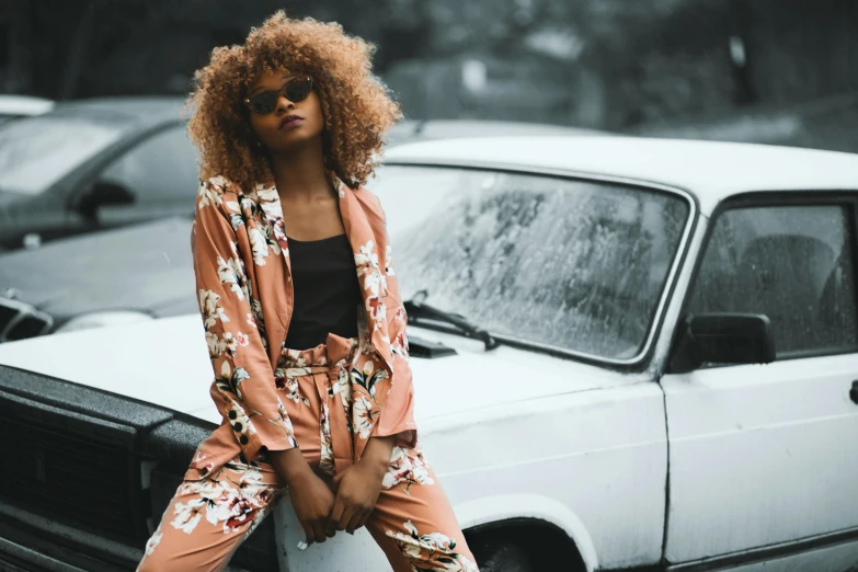 a woman sitting on top of a white car, a photo, trending on pexels, afrofuturism, floral clothes, wearing a worn out brown suit, curly haired, wearing a duster coat