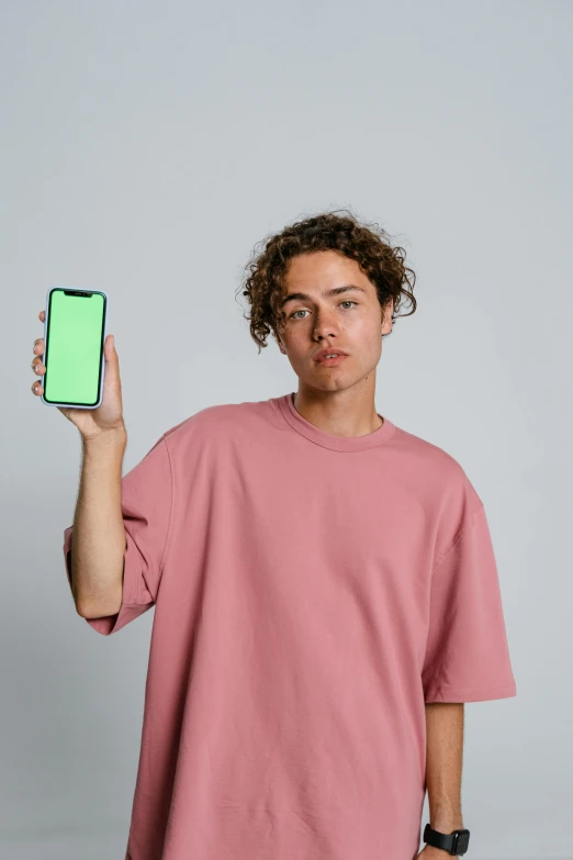 a man in a pink shirt holding up a green phone, trending on pexels, color field, joe keery, plain background, curly haired, trending on r/streetwear