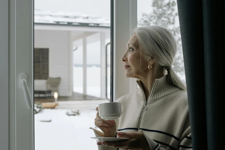 a woman holding a cup and looking out a window, inspired by Louisa Matthíasdóttir, pexels contest winner, high snow, gray haired, high resolution, outside winter landscape