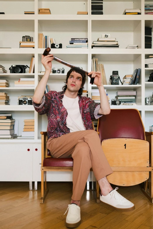 a man sitting on a chair in front of a bookshelf, an album cover, inspired by Davide Sasselli, trending on pexels, non binary model, mid - length hair, corduroy, h3h3