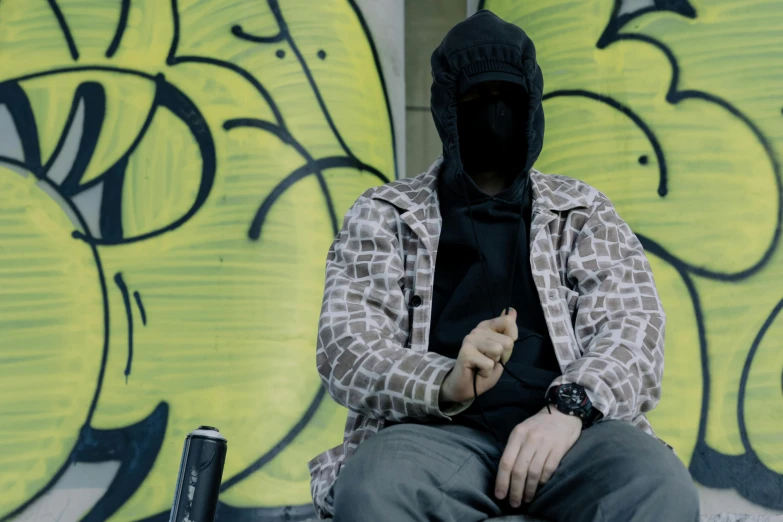 a man sitting in front of a graffiti covered wall, unsplash, graffiti, a hooded killer a cane, hands not visible, still image from tv series, balaclava