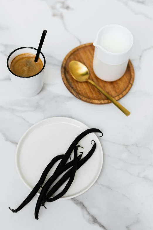 a white plate topped with vanilla beans next to a cup of coffee, a still life, trending on pexels, minimalism, white marble and gold, black fork, on kitchen table, umbra