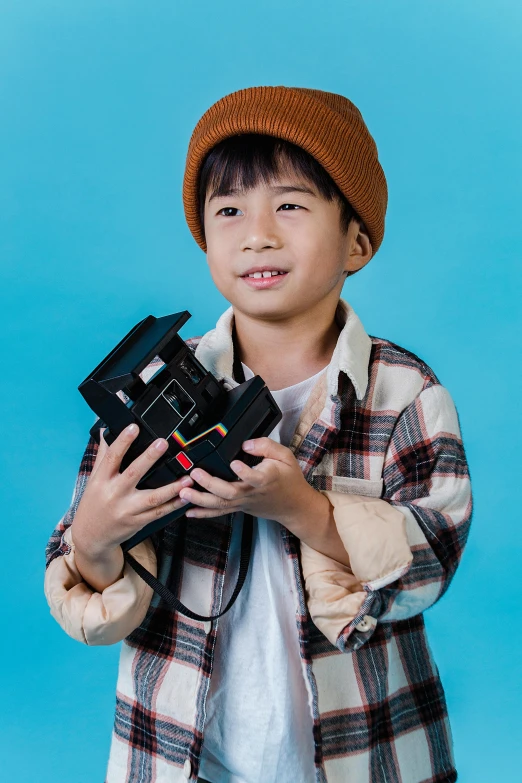 a young boy holding a camera on a blue background, inspired by Reuben Tam, pexels contest winner, caracter with brown hat, next gen, petite, slide show