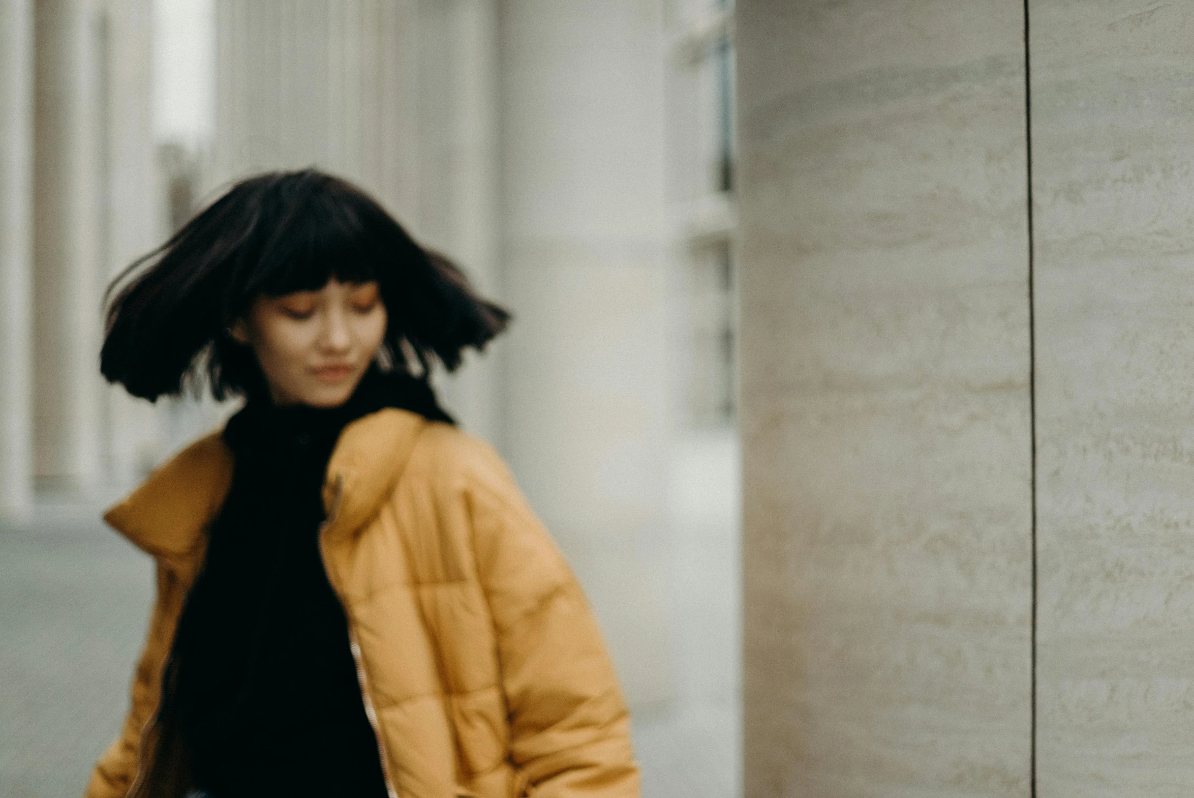 a woman in a yellow jacket standing next to a wall, trending on pexels, sui ishida with black hair, a woman walking, muted colors. ue 5, fluffy bangs
