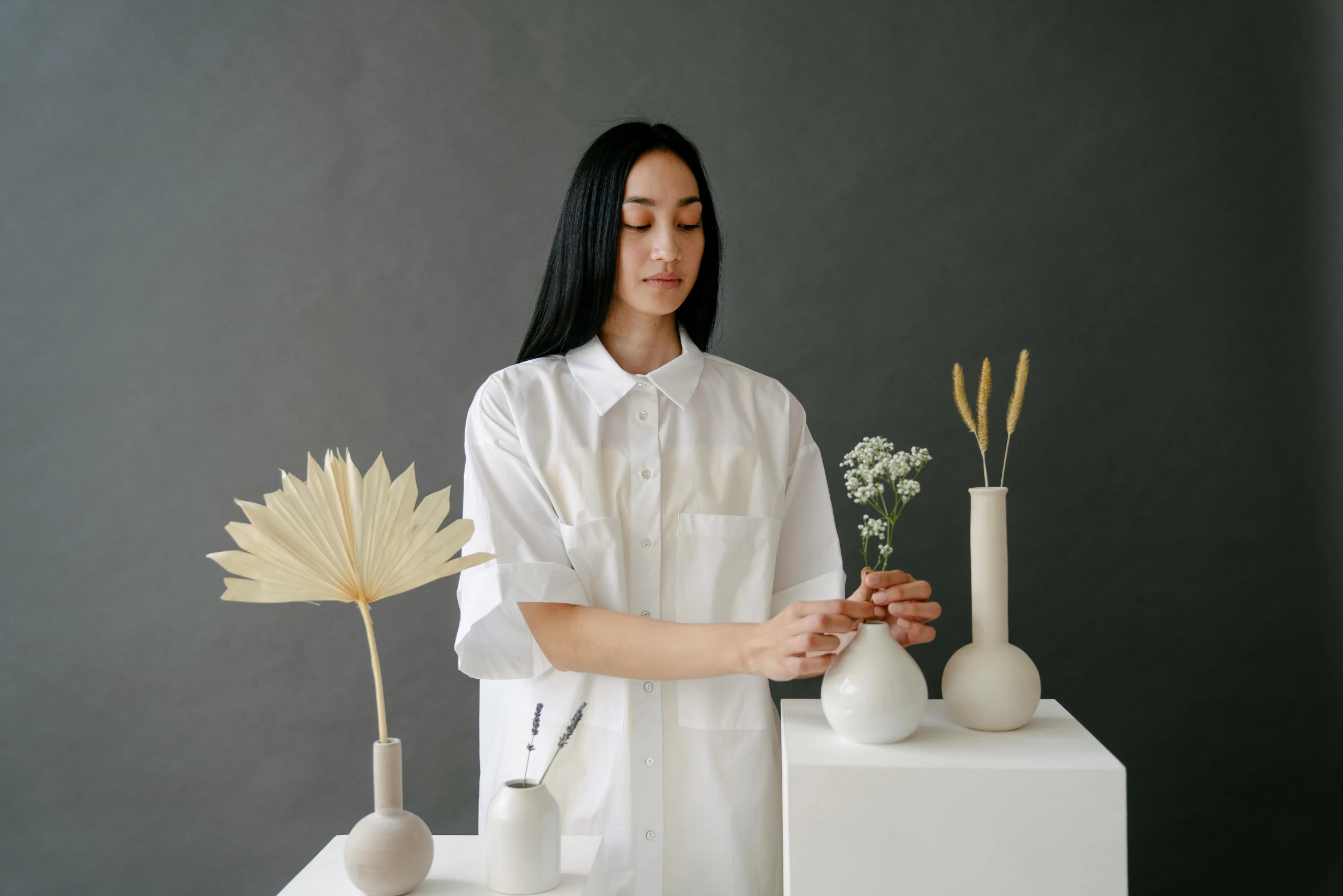 a woman standing in front of a bunch of vases, inspired by Kim Tschang Yeul, pexels contest winner, lab coat and tee shirt, wearing a white button up shirt, ikebana, detailed product image