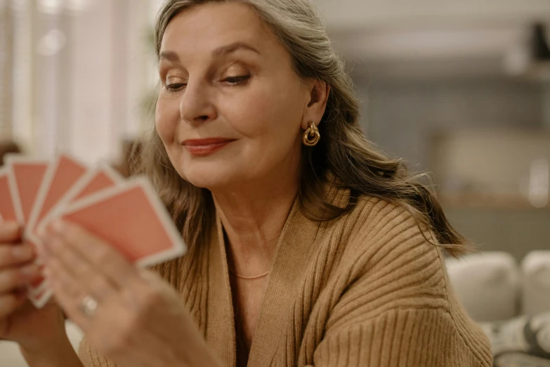 a woman sitting at a table holding playing cards, pexels contest winner, gray haired, blushing, youtube thumbnail, high quality image