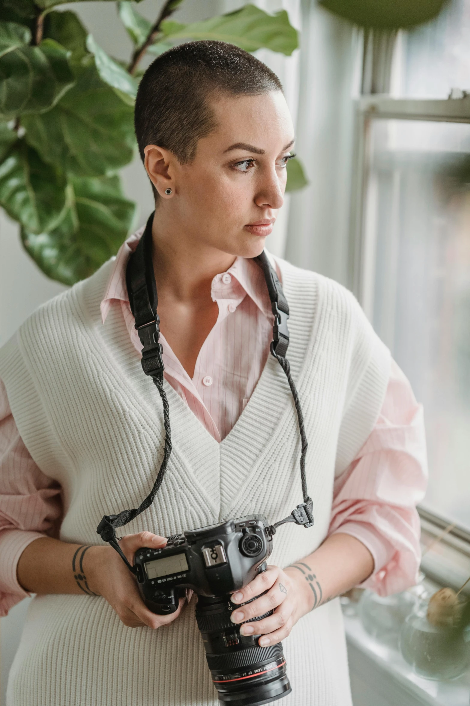 a woman holding a camera in front of a window, a portrait, by Winona Nelson, short shaved hair, pink body harness, wearing a white button up shirt, model is wearing techtical vest