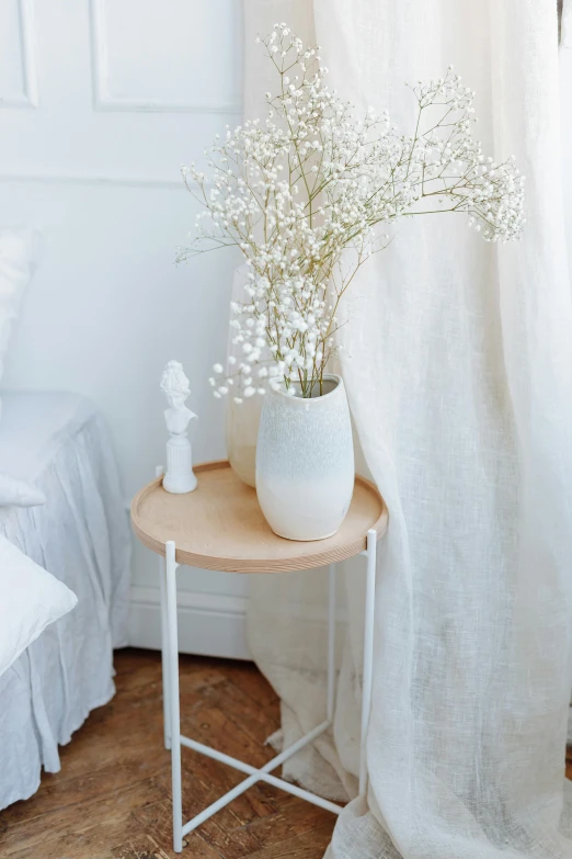 a white vase sitting on top of a table next to a bed, by Marie Angel, light and space, natural wood top, sheer fabrics, holding flowers, product photo
