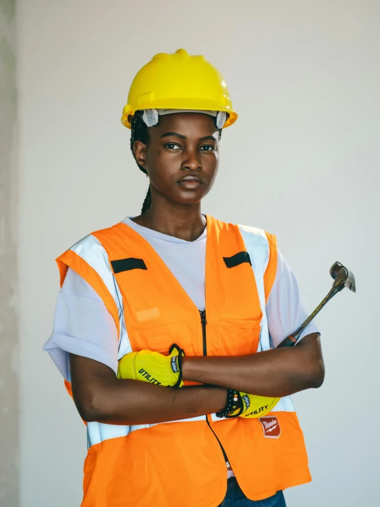 a woman in an orange safety vest holding a hammer, inspired by Afewerk Tekle, pexels contest winner, black young woman, yellow helmet, offwhite, ( ( dark skin ) )
