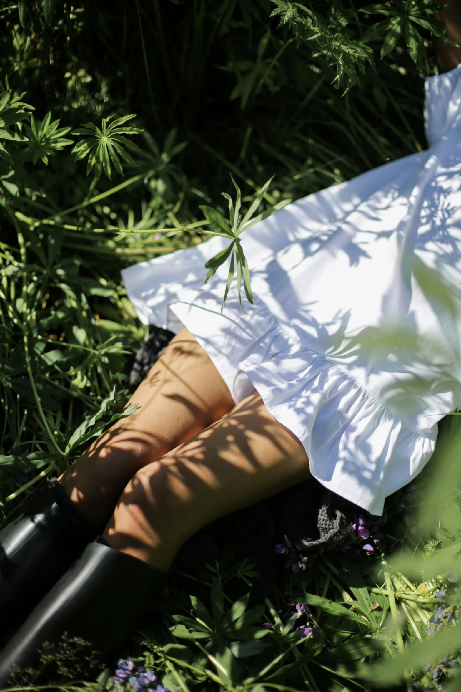 a woman laying on top of a lush green field, inspired by Andrew Wyeth, unsplash, white blouse and gothic boots, close-up on legs, sun and shadow, wearing a white folkdrakt dress