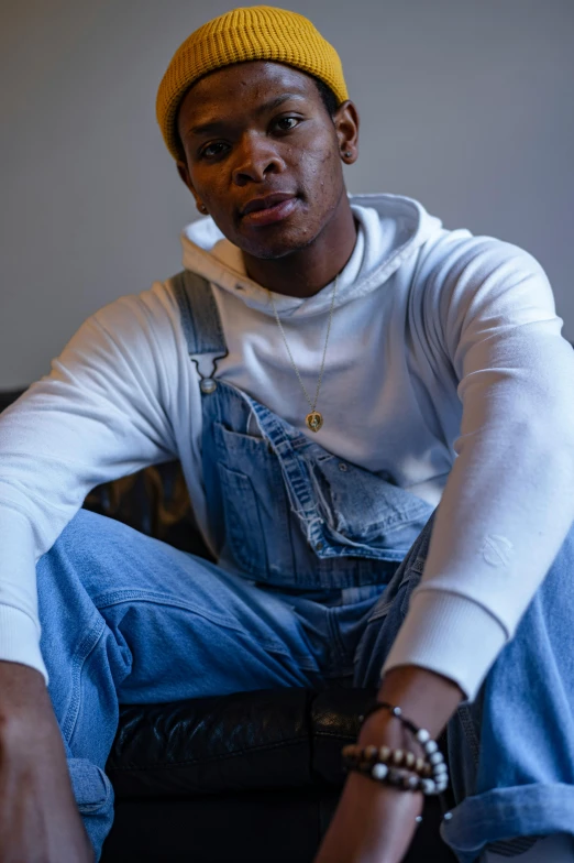 a man sitting on a couch wearing a yellow hat, inspired by Theo Constanté, trending on pexels, wearing blue jean overalls, wearing a white sweater, jemal shabazz, non binary model