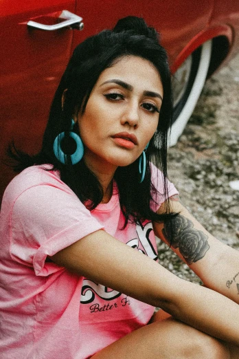a woman sitting on the ground next to a red car, an album cover, inspired by Elsa Bleda, trending on pexels, graffiti, beautiful iranian woman, closeup portrait shot, pink clothes, wearing a muscle tee shirt