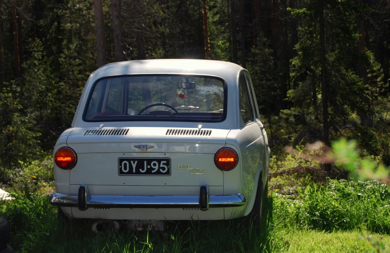 a small white car sitting on top of a lush green field, inspired by Veikko Törmänen, purism, pine forests, rear lighting, classic cars, year 1968