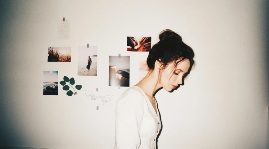 a woman standing in front of a wall covered in pictures, a polaroid photo, inspired by Elsa Bleda, trending on pexels, aestheticism, girl with messy bun hairstyle, side profile artwork, porcelain pale skin, music album cover