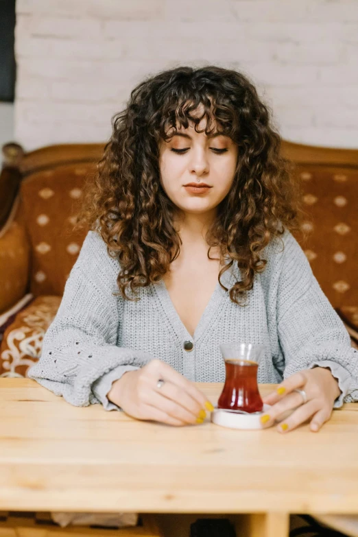 a woman sitting at a table with a cup of tea, a portrait, by Julia Pishtar, trending on pexels, drinking cough syrup, curly bangs, young middle eastern woman, gif