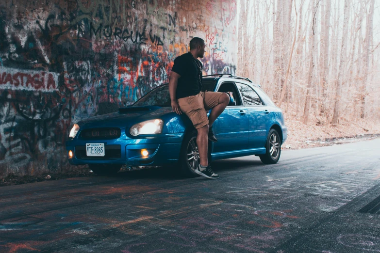 a man standing on the hood of a blue car, by Marshall Arisman, pexels contest winner, graffiti, subaru, sitting down casually, teddy fresh, 2000s photo