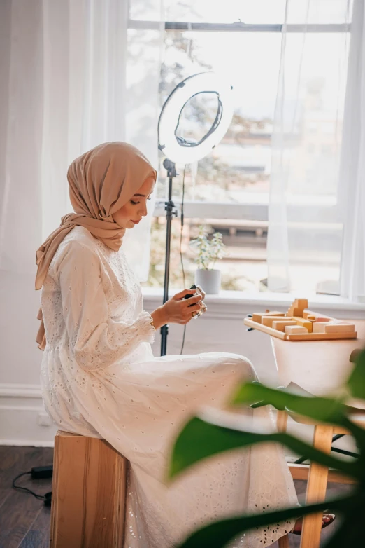 a woman sitting on a stool looking at her phone, a picture, inspired by Maryam Hashemi, trending on pexels, hurufiyya, white hijab, photoshoot for skincare brand, ring light, crafting