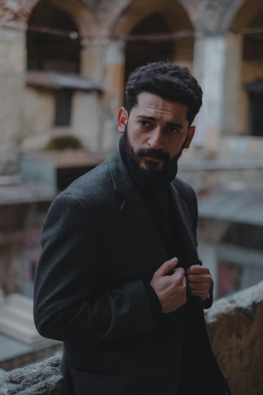 a man in a black coat standing on a balcony, an album cover, inspired by Carlo Mense, pexels contest winner, bearded beautiful man, real life photo of a syrian man, ( ( theatrical ) ), vintage color