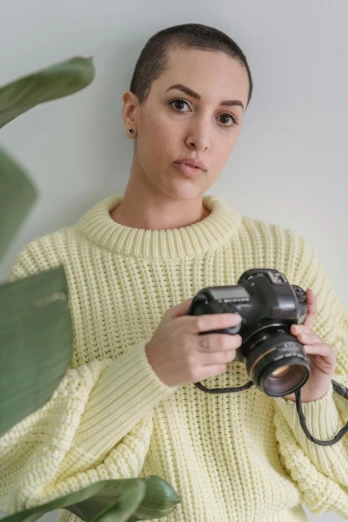 a woman holding a camera in front of a plant, wearing casual sweater, modern studio light soft colour, bella poarch, on a yellow canva