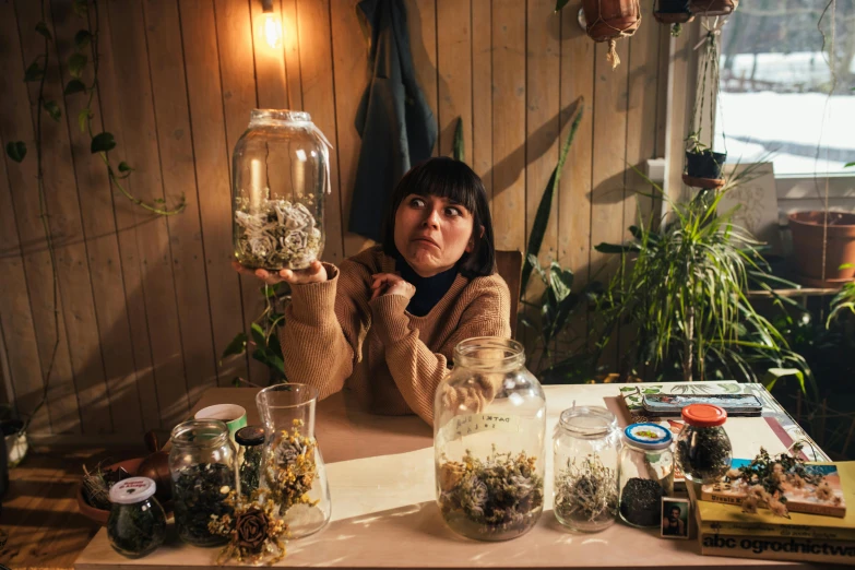 a woman sitting at a table holding a jar of plants, process art, glass domes, charli bowater and artgeem, sprawling, biophilia mood