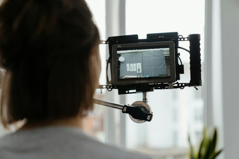 a woman holding a video camera in front of a window, by Matthias Stom, unsplash, video art, looking at monitor, with a robotic arm, close-up shot taken from behind, behind the scenes photo