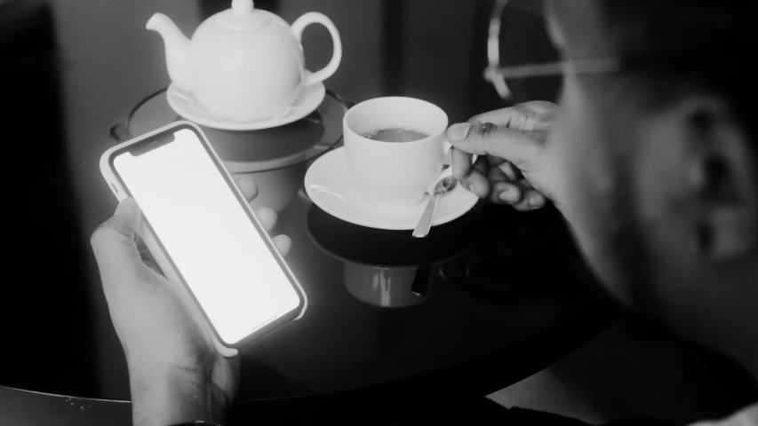 a person sitting at a table with a cell phone, a black and white photo, by Emma Andijewska, pexels, tea cup, avatar image, square, thumbnail