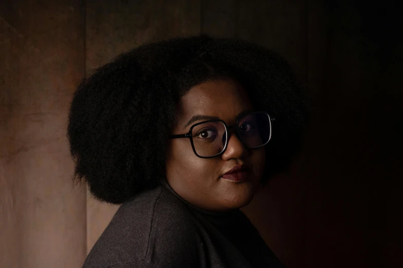 a woman with glasses standing in front of a wooden wall, by Lily Delissa Joseph, dark skin, portrait image, a portrait of a plump woman, studio shot