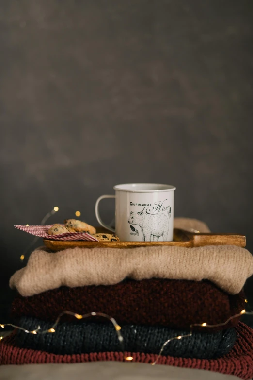 a cup of coffee sitting on top of a pile of sweaters, a portrait, inspired by Muggur, hurufiyya, dark grey backdrop studio, on a wooden tray, full product shot, cozy candlelight