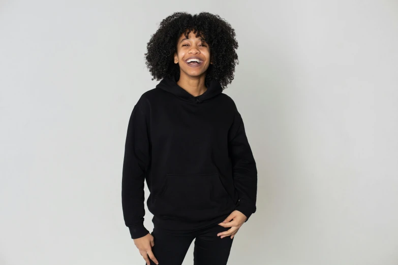 a woman standing in front of a white wall wearing a black hoodie, smiling, all black matte product, full color, f / 2 0