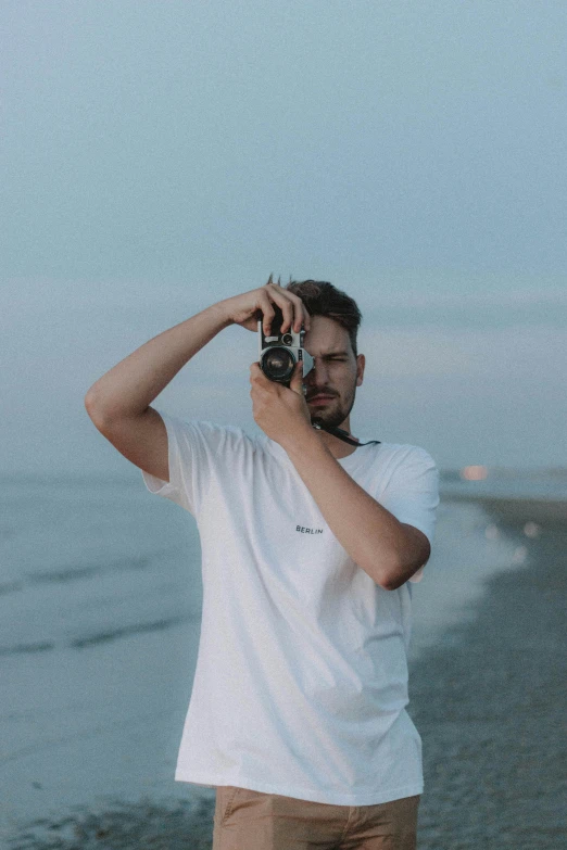 a man standing on a beach holding a camera, dressed in a white t shirt, 2019 trending photo, profile picture, liam