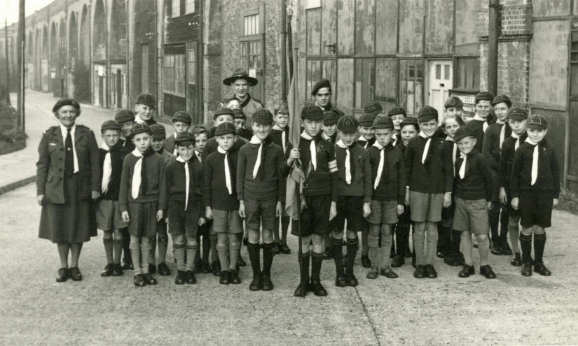 a group of children standing in front of a building, a black and white photo, by Sylvia Wishart, scout boy, each wearing correct era clothes, kings row in the background, in formation