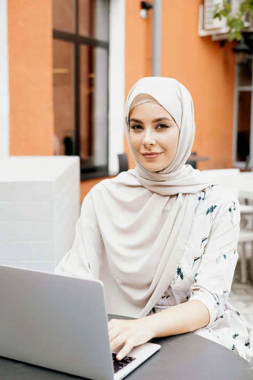 a woman sitting at a table with a laptop, inspired by Maryam Hashemi, trending on pexels, hurufiyya, wearing robe, malaysian, confident smirk, white scarf