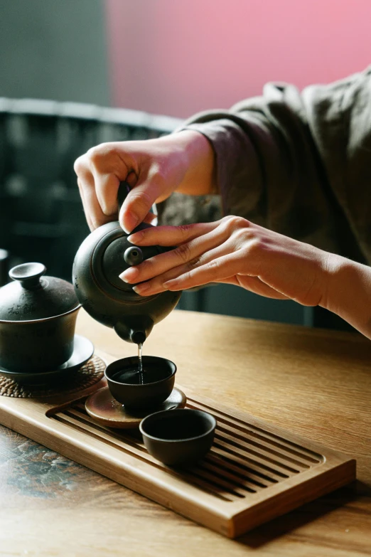 a person pouring tea into a cup on a wooden tray, inspired by Shūbun Tenshō, trending on unsplash, square, wearing black robe, teapots, 15081959 21121991 01012000 4k