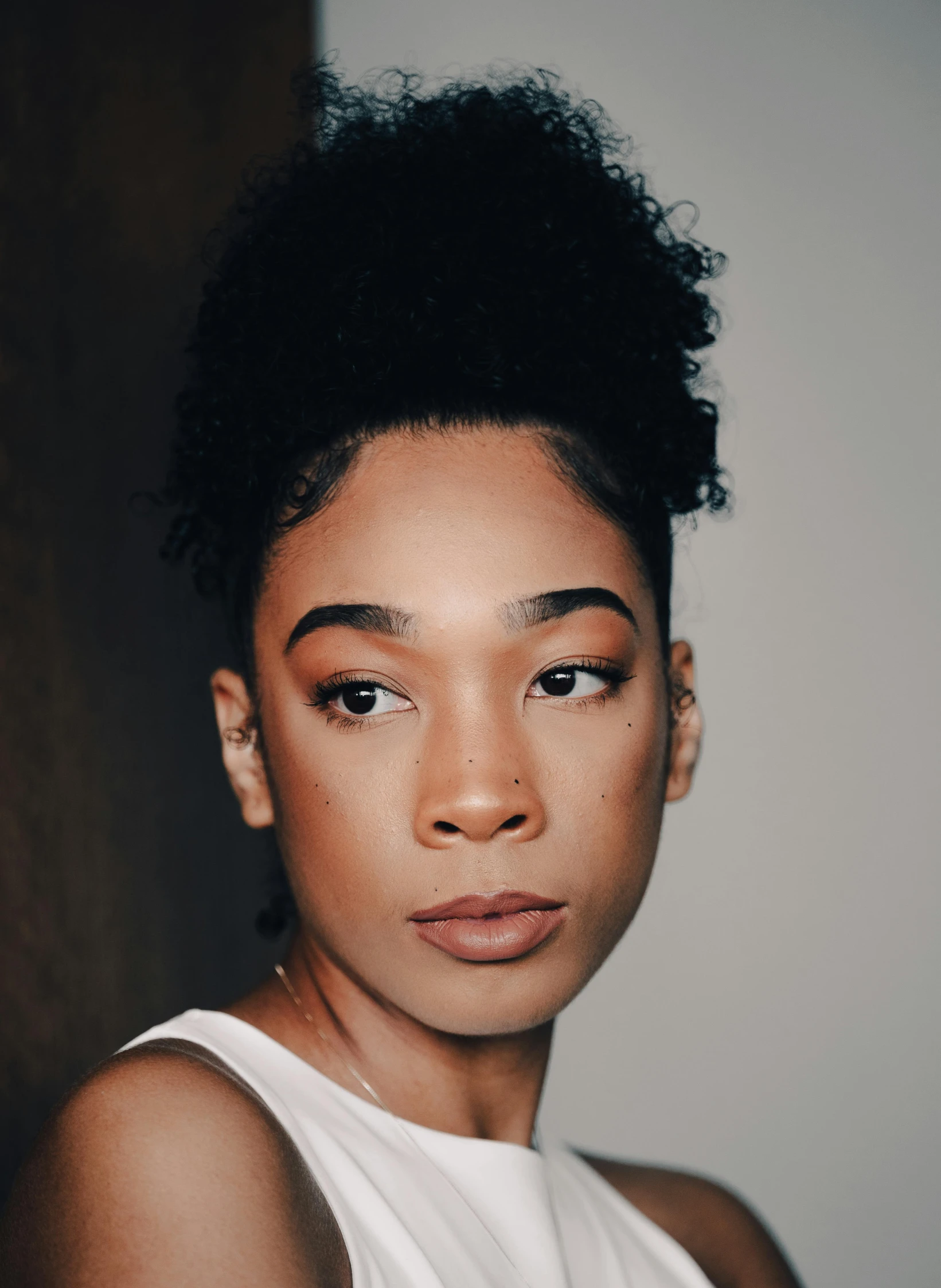 a woman with black hair wearing a white tank top, by Lily Delissa Joseph, trending on pexels, hyperrealism, curly messy high bun hairstyle, ☁🌪🌙👩🏾, androgynous face, ashteroth