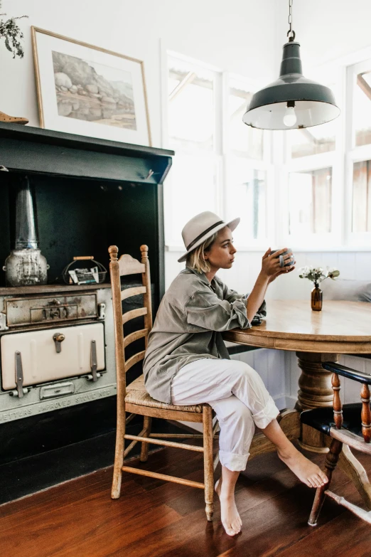 a woman sitting at a table in a room, by Elsie Henderson, pexels contest winner, wearing a travel hat, cottage decor, coffee, white