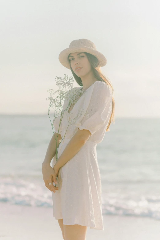a woman standing on a beach holding a bunch of flowers, unsplash, renaissance, white suit and hat, portrait of ana de armas, thoughtful ), sunfaded