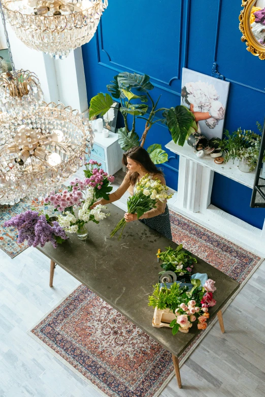 a woman arranging flowers on a table in a room, maximalism, glowing blue interior components, ariel view, curated collections, carrying flowers