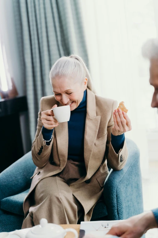 a couple of people that are sitting in a chair, drinking a cup of coffee, older woman, taking control while smiling, snacks