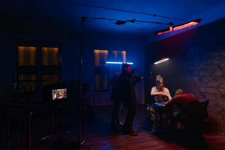 a group of people sitting around a table in a dark room, inspired by Nan Goldin, video art, blue and red lights, movie setup, behind the scenes photo, vhs footage of a movie set