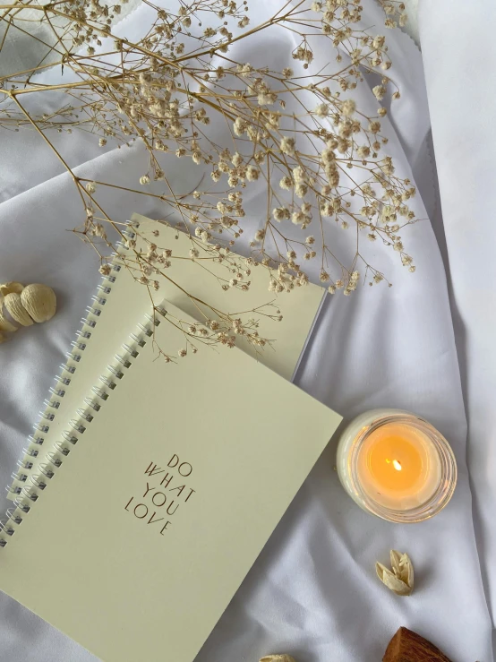 a book sitting on top of a bed next to a candle, white and yellow scheme, holding notebook, promo image, noot noot