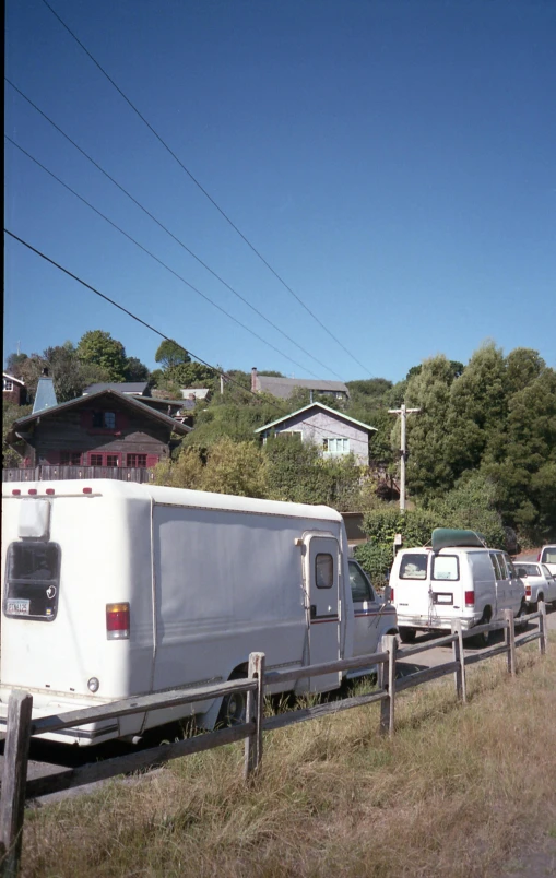 a long line of cars parked on the side of the road, by Jim Nelson, ca. 2001, built on a steep hill, small houses, view from a news truck