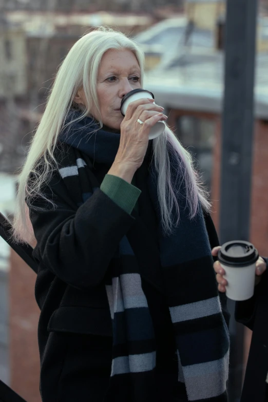 a woman drinking a cup of coffee on top of a building, a photo, inspired by Louisa Matthíasdóttir, trending on pexels, photorealism, long grey hair, wearing long black winter coat, people walking around, cinematic shot ar 9:16 -n 6 -g