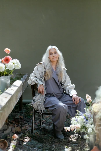 a woman sitting in a chair surrounded by flowers, a portrait, inspired by Grethe Jürgens, unsplash, light gray long hair, artist wearing overalls, wearing long silver robes, sitting on a mocha-colored table